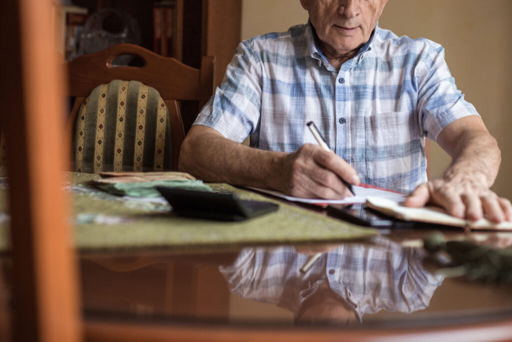 A senior man is sitting at a dining room table paying bills. He has a calculator in front of him.