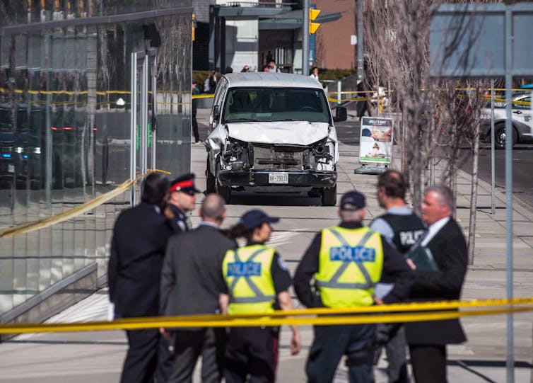 Police stand on a sidewalk in front of the damaged white van